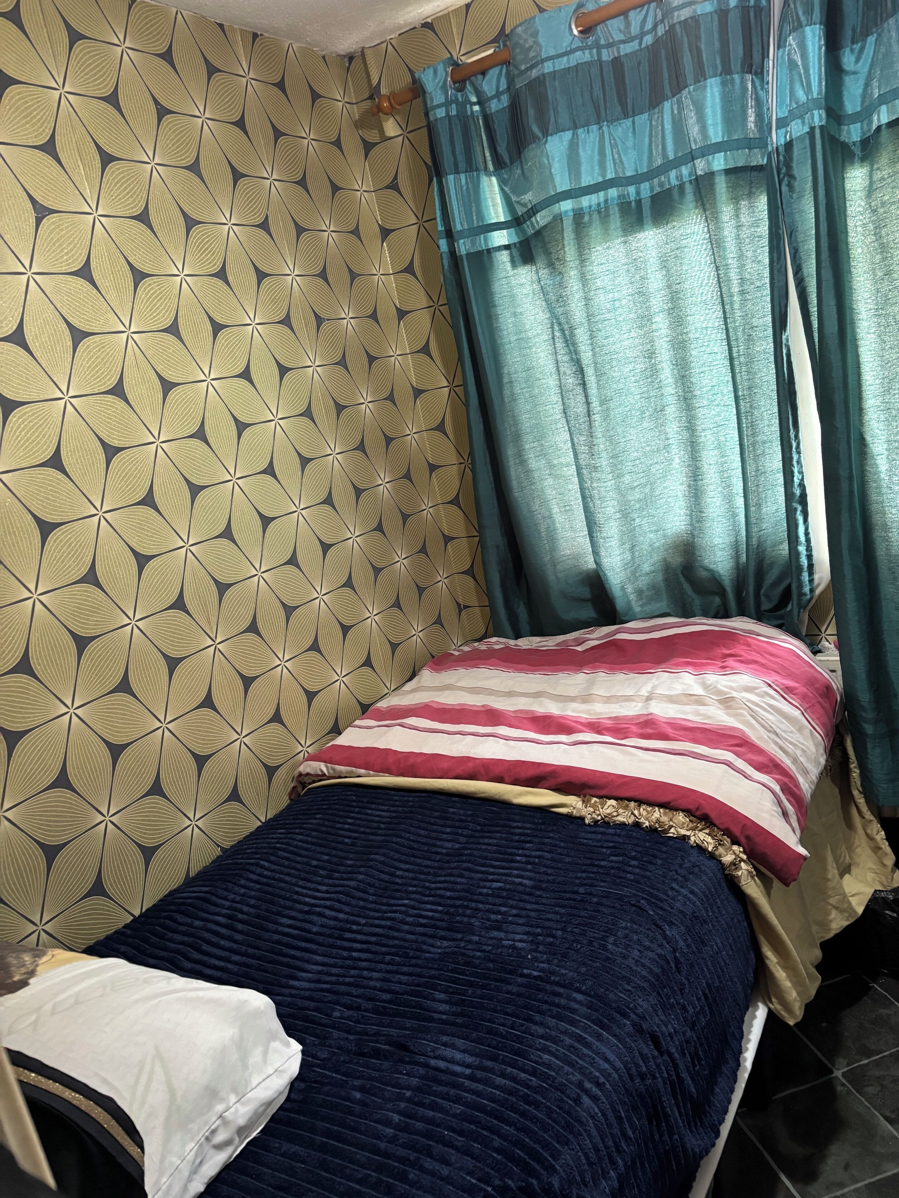 Una habitación pequeña con cama individual, papel tapiz estampado, colcha a rayas y cortinas color turquesa.