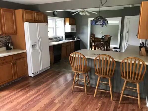 Una cocina con armarios de madera, un refrigerador blanco, una zona de comedor con sillas de madera y un ventilador de techo.