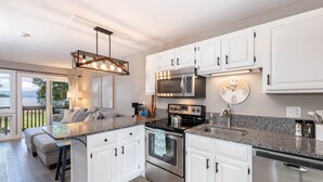 Une cuisine moderne avec des meubles blancs, des appareils en acier inoxydable, un comptoir en granit et une grande fenêtre donnant sur une terrasse.