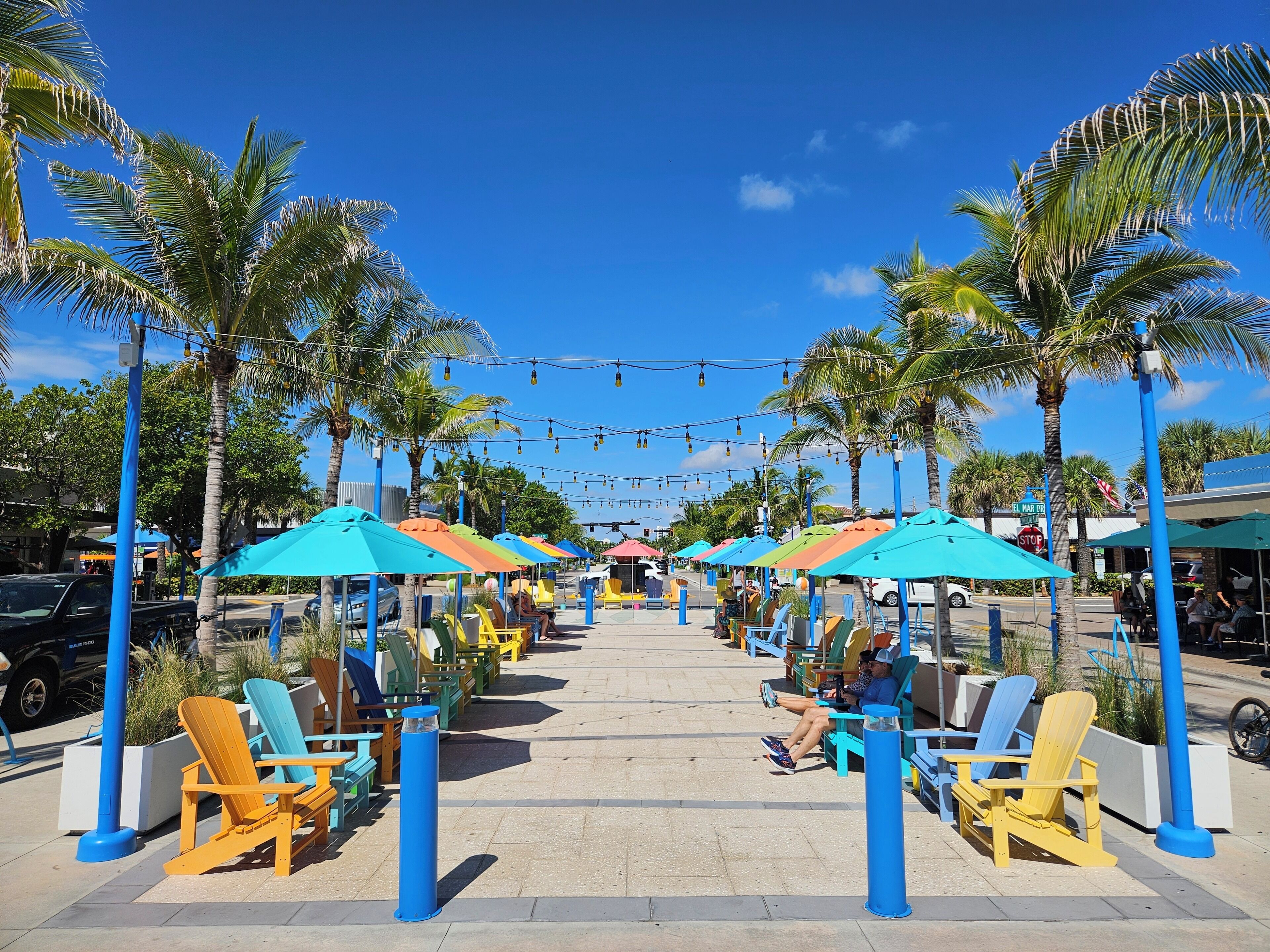 A beachside seating area with colorful umbrellas and chairs, palm trees, and a clear blue sky.