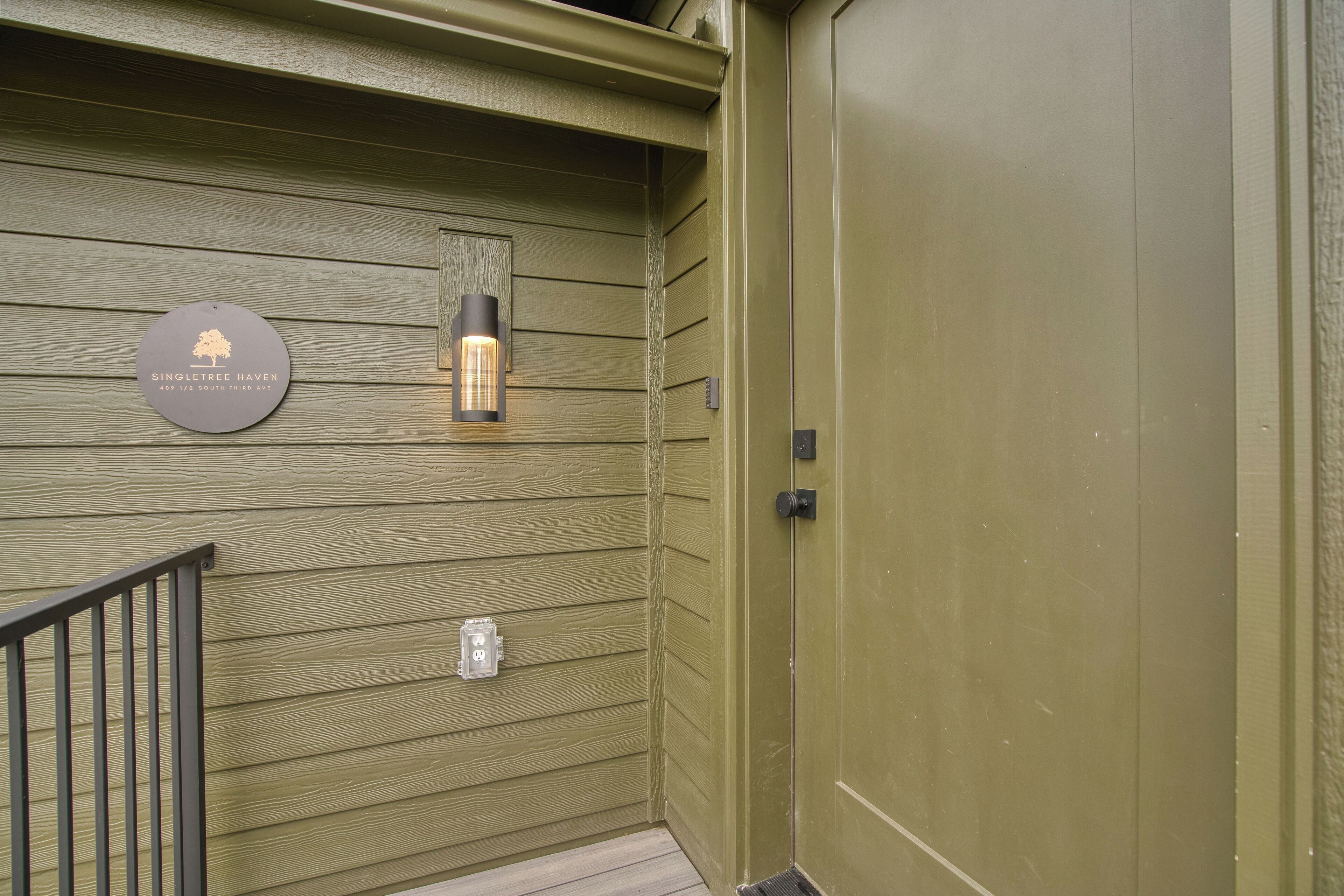 Pared exterior de madera con una puerta de metal, un accesorio de luz y un letrero con el logotipo de un árbol.