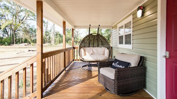 A porch with a wicker chair and a cushioned bench, a wooden deck, and a red door. - Lovely cozy cottage with AC in charming Waycross (Waycross)