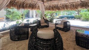 Un'area salotto esterna con sedie e tavoli in rattan, un tetto di paglia e vista sul verde circostante.