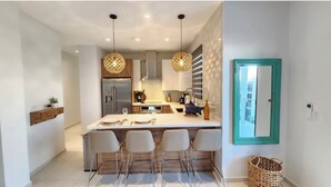 Una cucina moderna con isola centrale, sgabelli da bar, luci a sospensione e una porta con cornice color petrolio.