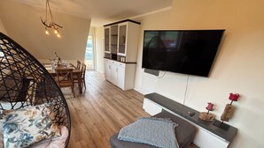 Een moderne woonkamer met een eethoek, een grote flatscreen televisie aan de muur en een gezellige zithoek met een gestoffeerde bank en een rieten stoel.