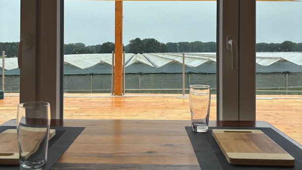 Een eethoek met uitzicht op kassen en een houten tafel met twee glazen en een houten plank.