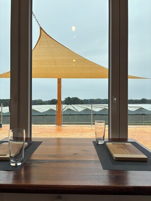 Comedor con vistas a invernaderos y una mesa de madera con dos vasos y una tabla de madera.