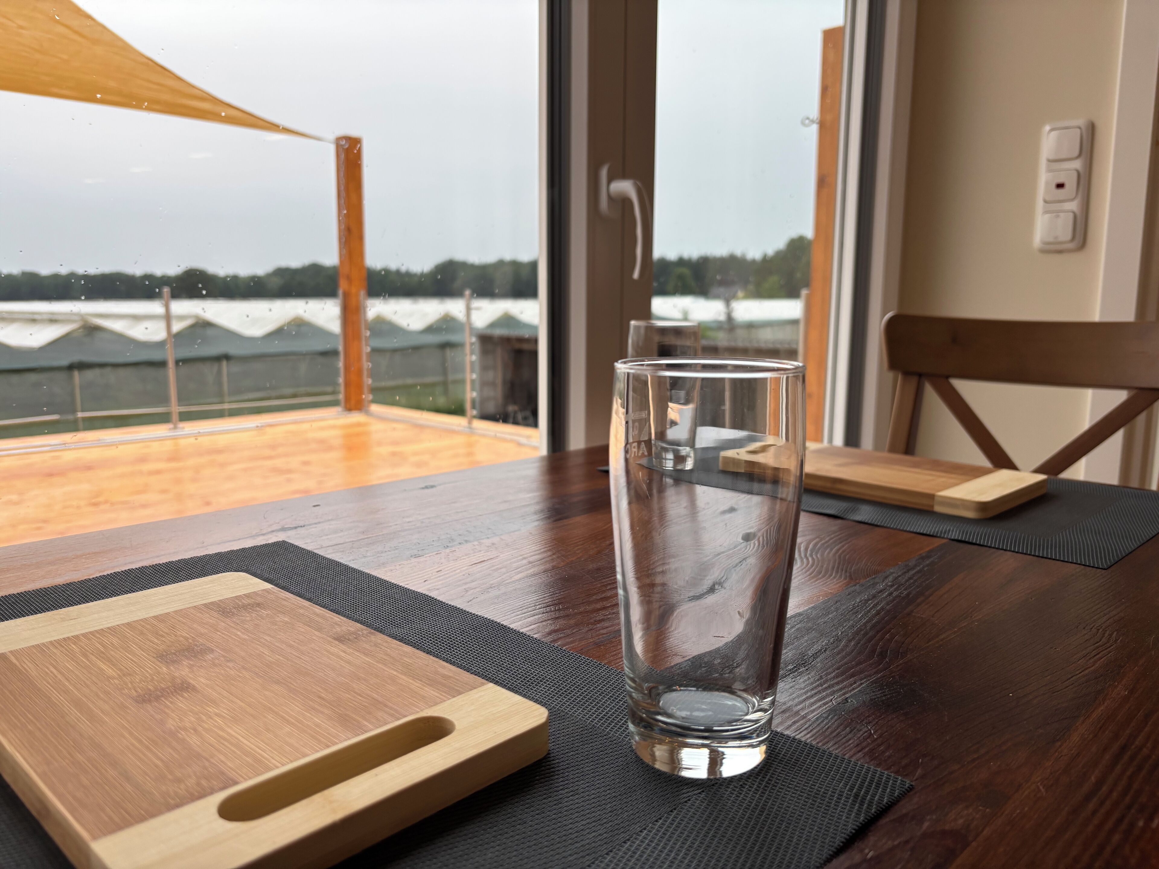 A glass of water on a wooden table with a view of a green field through a window.