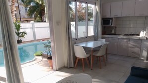 Una cocina moderna con armarios blancos, un comedor con mesa y sillas blancas, y vista a una piscina exterior.