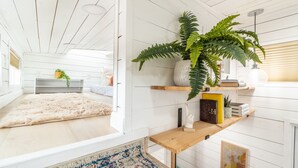 A modern, well-lit room with a large window, a cozy rug, a wooden shelf with decorative items, and a potted plant.
