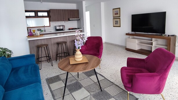 A modern living room with a kitchen in the background, a flat-screen TV, and vibrant chairs. - Central and spacious apartment in Rionegro (Rionegro)
