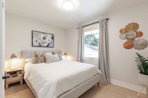 Un dormitorio con cama, mesitas de noche, lámpara, ventana con cortinas y adornos en la pared.