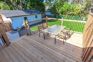 Una terraza de madera con una mesa y sillas de vidrio, rodeada por una valla metálica y un cobertizo pequeño.