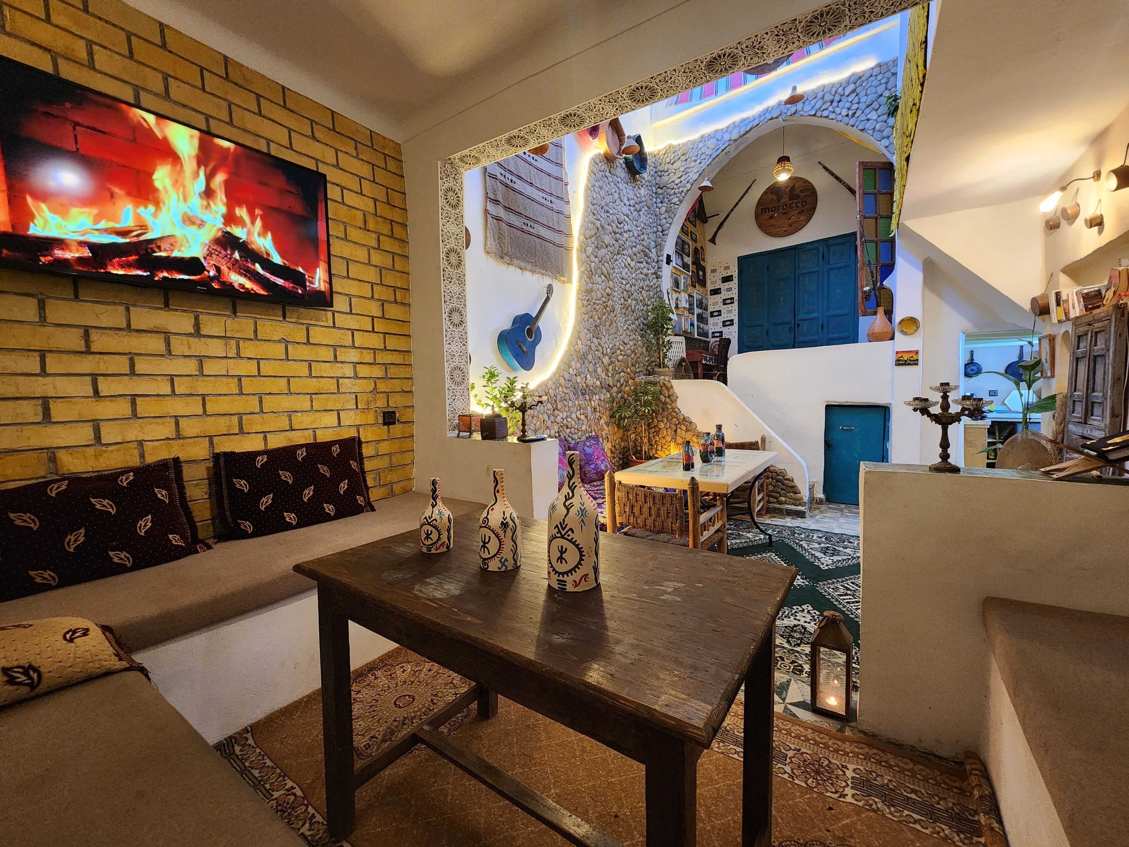 A cozy living area with a brick wall, a flat-screen TV, a wooden table with decorative vases, and a blue door.
