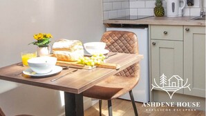 La cuisine est équipée d’un four à micro-ondes, d’un grille-pain et d’une machine à café. On y trouve une table avec une miche de pain, un saladier de fruits et un verre de jus d’orange.