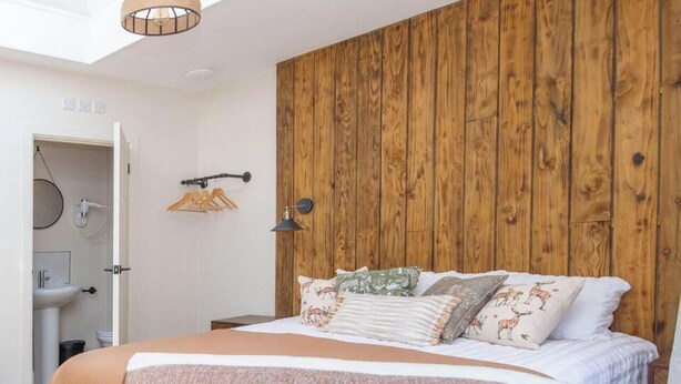 Une chambre avec un mur lambrissé en bois, un lit avec de nombreux oreillers, un luminaire suspendu et une salle de bain visible avec un lavabo.