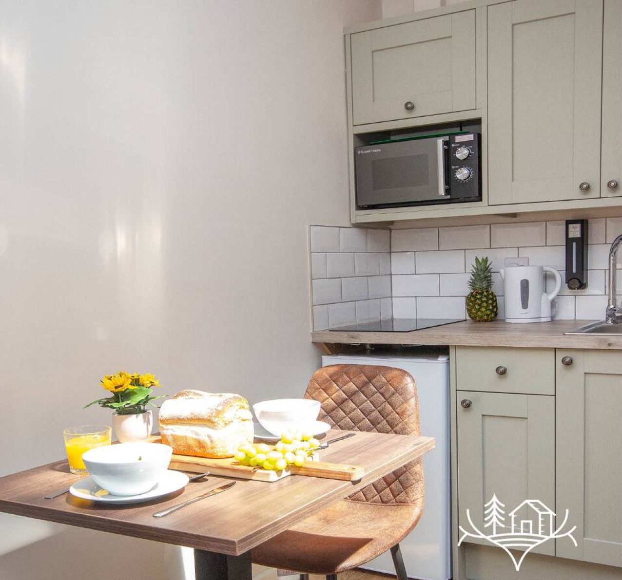 La cuisine est équipée d’un four à micro-ondes, d’un grille-pain et d’une machine à café. On y trouve une table avec une miche de pain, un saladier de fruits et un verre de jus d’orange.