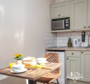 A kitchen with a microwave, a toaster, and a coffee maker. There is a table with a loaf of bread, a bowl of fruit, and a glass of orange juice. - The Noble Stag - Room 3 - New Forest (Dibden Purlieu)