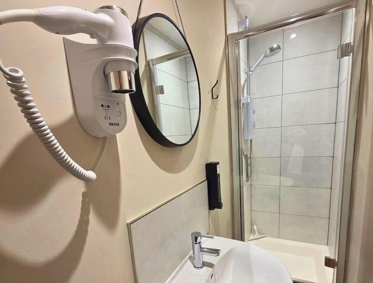 Une salle de bain avec un lavabo blanc, un sèche-cheveux, un miroir et un espace douche.
