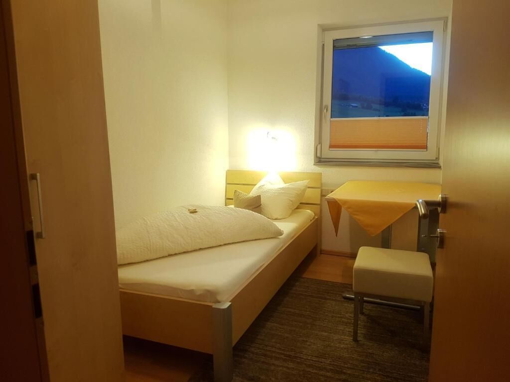 Une petite chambre bien éclairée, avec un lit simple, une table de chevet, une chaise et une fenêtre donnant sur une montagne.