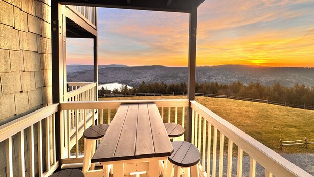 Un terrazzo in legno con tavolo e sedie affacciato su un panorama mozzafiato di montagne e un tramonto.