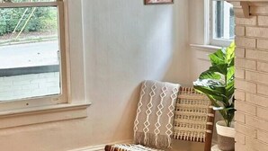 Quarto de canto com piso de madeira, uma cadeira de fibra natural, uma planta em vaso e quadros nas paredes.