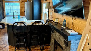 A cozy kitchen with wooden walls, a large TV, a refrigerator, a sink, and a bar area with chairs.