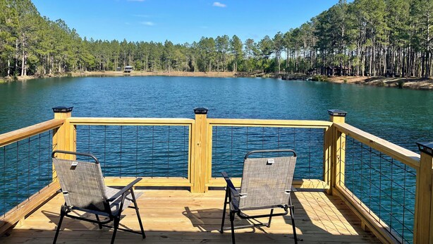 A wooden deck with two chairs overlooking a calm lake, surrounded by dense forest.