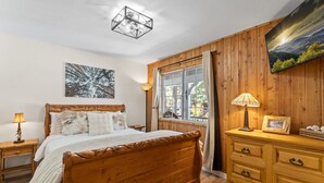 Ein Bett mit Holzkopfteil, eine Nachttischlampe, ein Nachttisch, eine Kommode mit Schubladen, ein Fenster mit Vorhängen und ein Bild an der Wand.