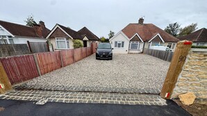 A residential driveway with a gravel surface leading to a black car parked in front of a house with a red-tiled roof.