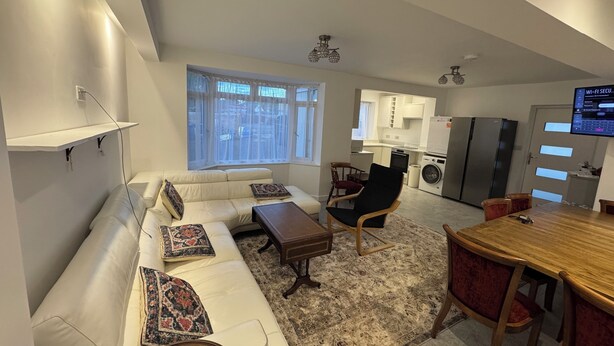 A modern living room with a white sofa, a wooden coffee table, and a dining table with chairs. There is a kitchen area with white cabinets and a refrigerator.
