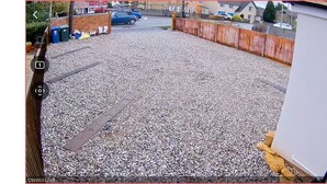 A gravel parking area with a metal grate, a wooden fence, and a blue car in the background.