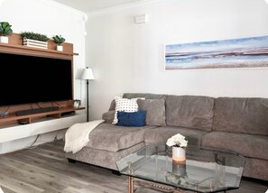 A living room with a grey sofa, a glass coffee table with a flower vase, a flat-screen TV on a wooden stand, and a painting on the wall. - 3-bedroom house Near everything in New Orleans (New Orleans)
