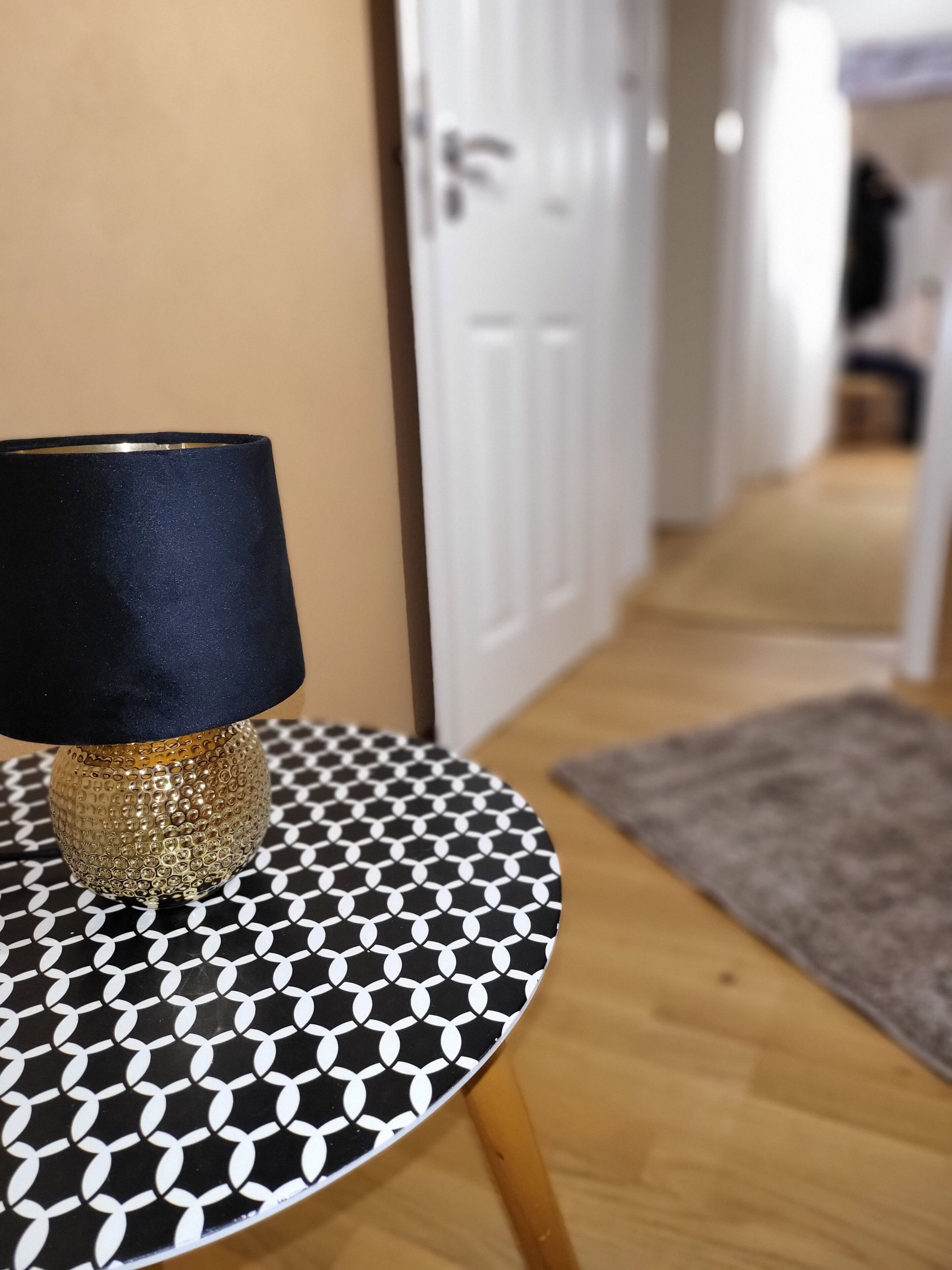 Ein runder Tisch mit einer schwarz-weißen Tischplatte und goldfarbenem Fuß, platziert in einer Diele mit Holzboden und grauem Teppich.