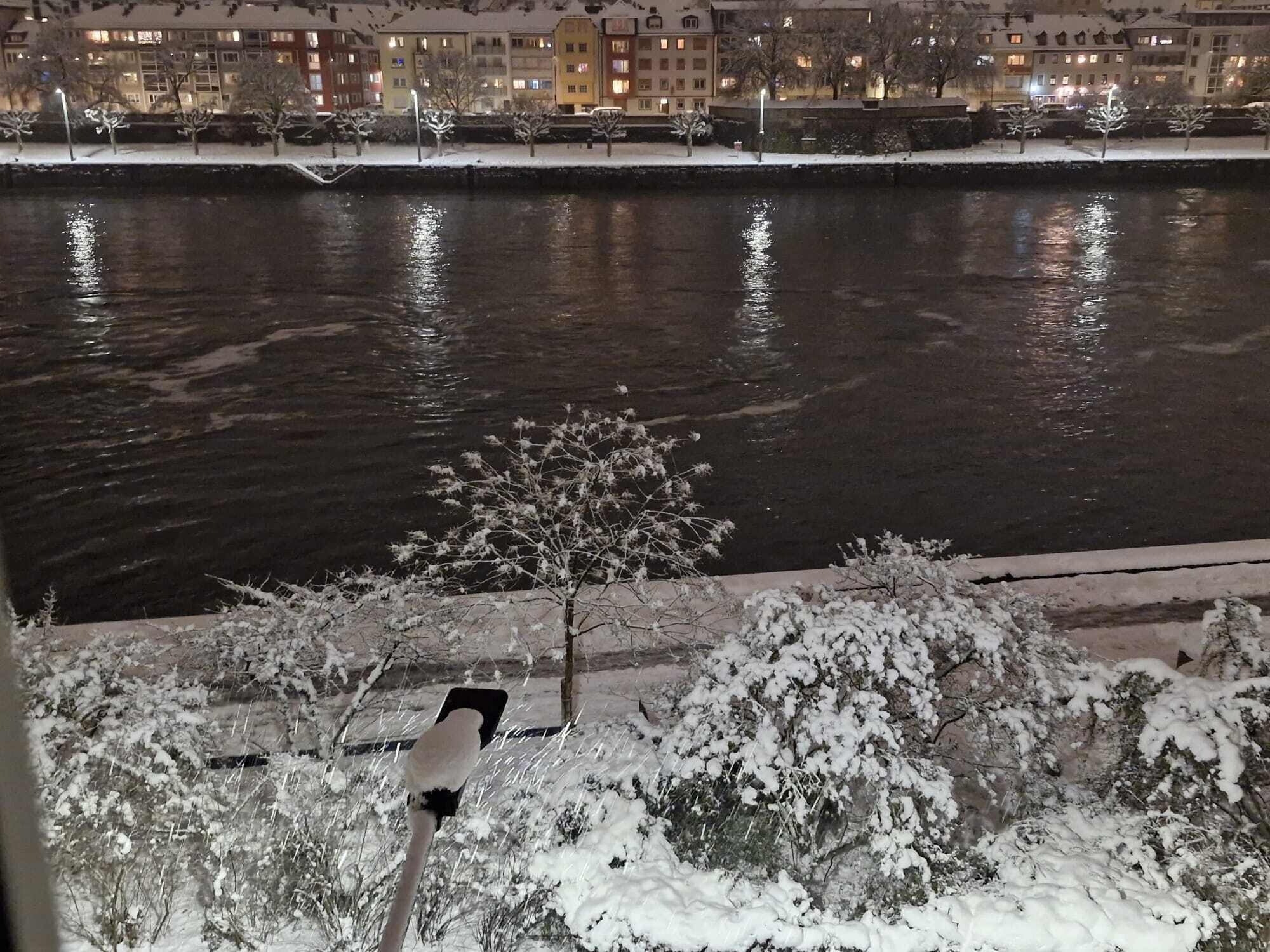 A nighttime view of a river with snow-covered trees and buildings in the background.