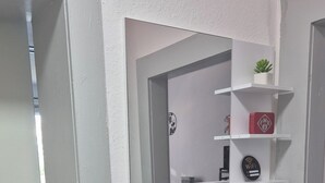 Ein schmaler Flur mit einer weißen Tür links. Ein roter Lanyard hängt am Türknauf, und oberhalb der Tür befindet sich eine kleine Topfpflanze auf einem weißen Regal.