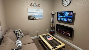 A living room with a beige sofa, a glass coffee table, a ceiling fan, a wall clock, and a flat-screen TV mounted on the wall.