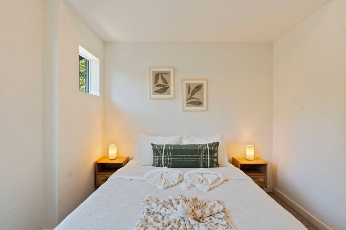 A neatly made bed with two pillows, a blanket, and towels folded on it. There are two bedside tables with lamps, and two framed pictures on the wall above the bed.