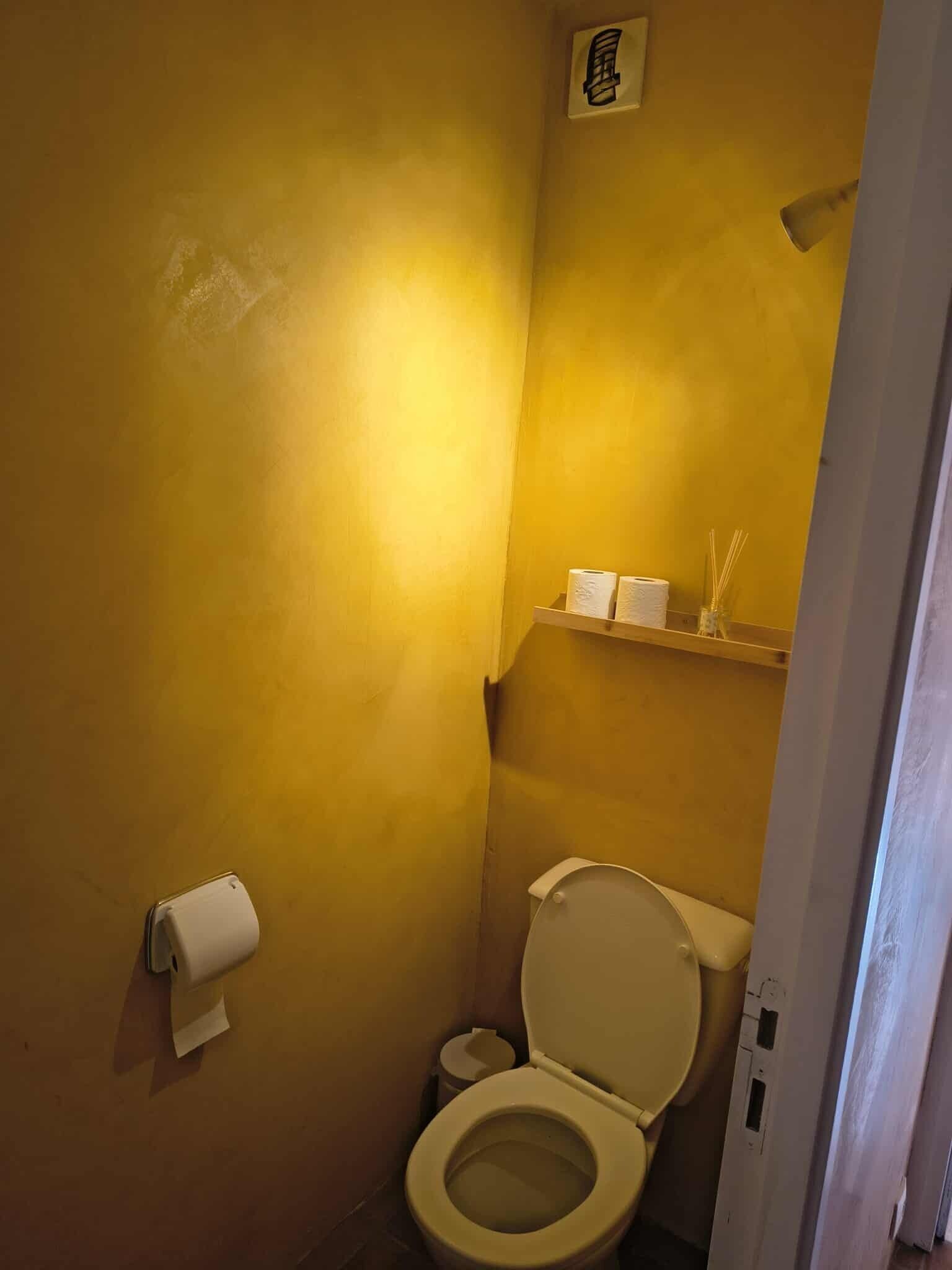 Un baño pequeño con paredes amarillas, un inodoro, un dispensador de toallas de papel y una repisa con artículos de aseo.