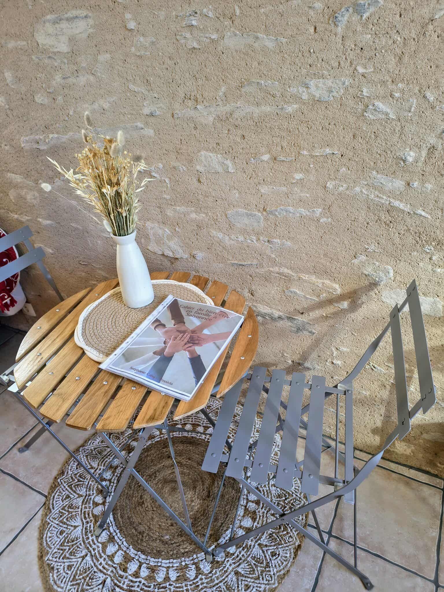 A round wooden table with a white vase of dried flowers and a magazine. A metal chair with a woven seat cushion is placed beside the table.