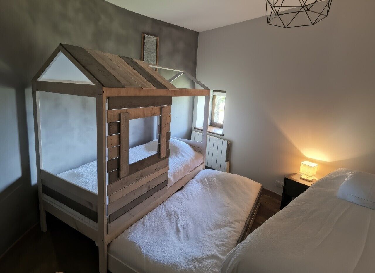 A bedroom with a wooden bunk bed, a bedside table with a lamp, and a window.