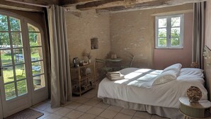 A rustic bedroom with a bed, a small table, a chair, a window, and a door leading to an outdoor area. - Saisonnée (Centrès)