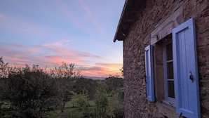 A rustic stone building with a wooden staircase leading to a balcony overlooking a pond and trees. - Saisonnée (Centrès)