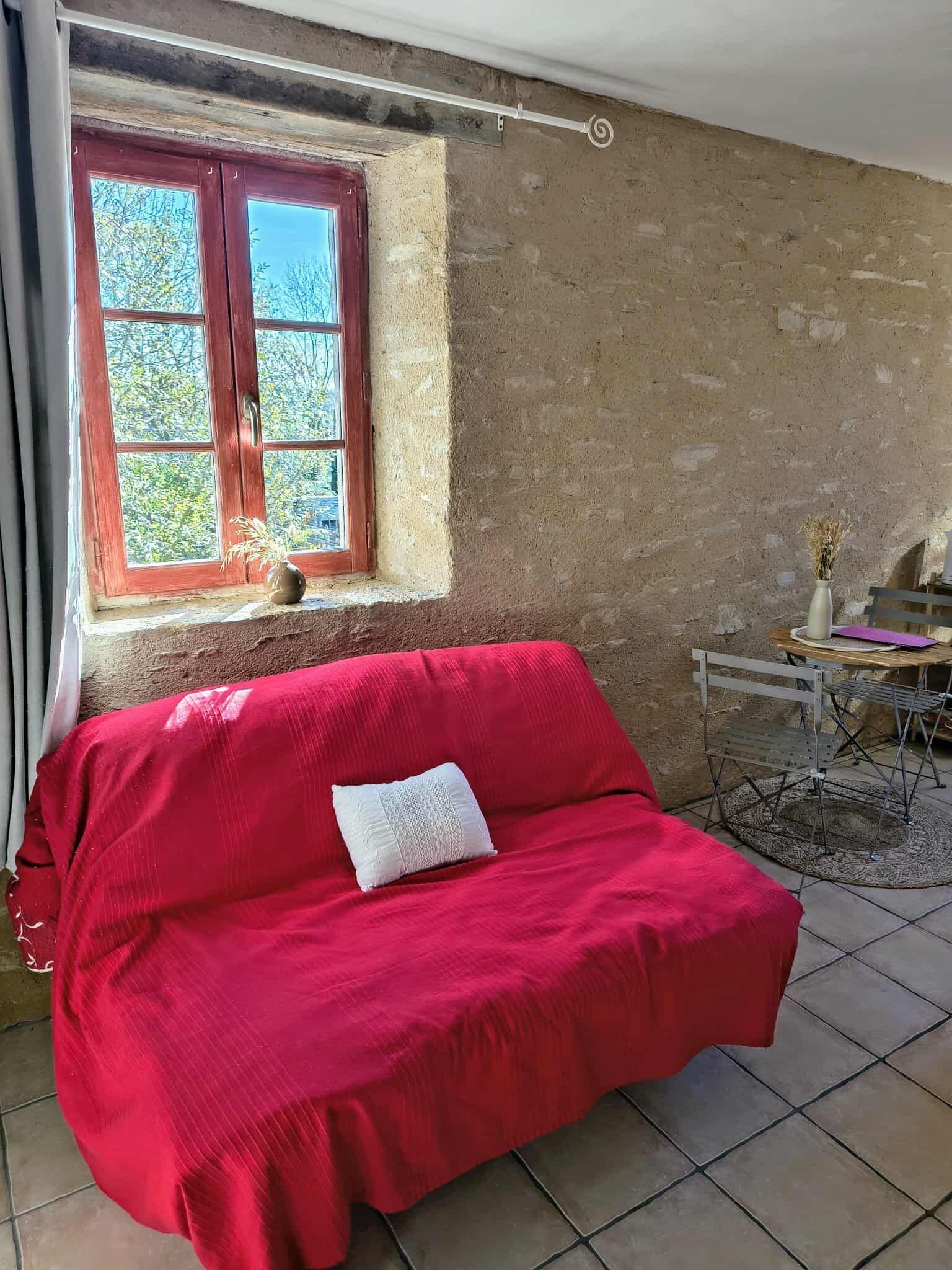 A room with a red sofa, a window with a view of trees, a table with a vase, and a chair.