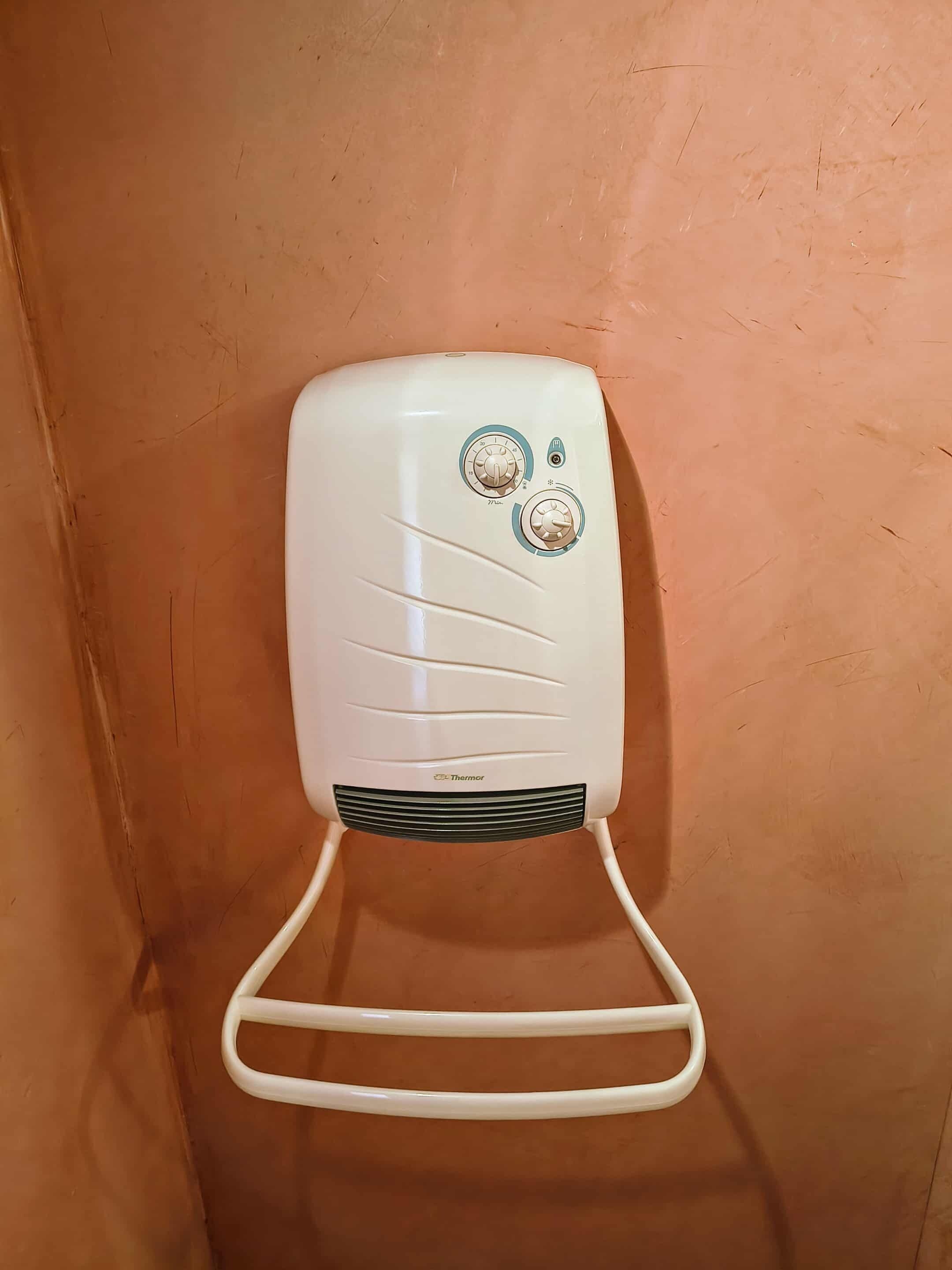 Calentador portátil blanco con dos mandos de control y el nombre de la marca en la parte frontal, apoyado sobre una pared naranja con textura.