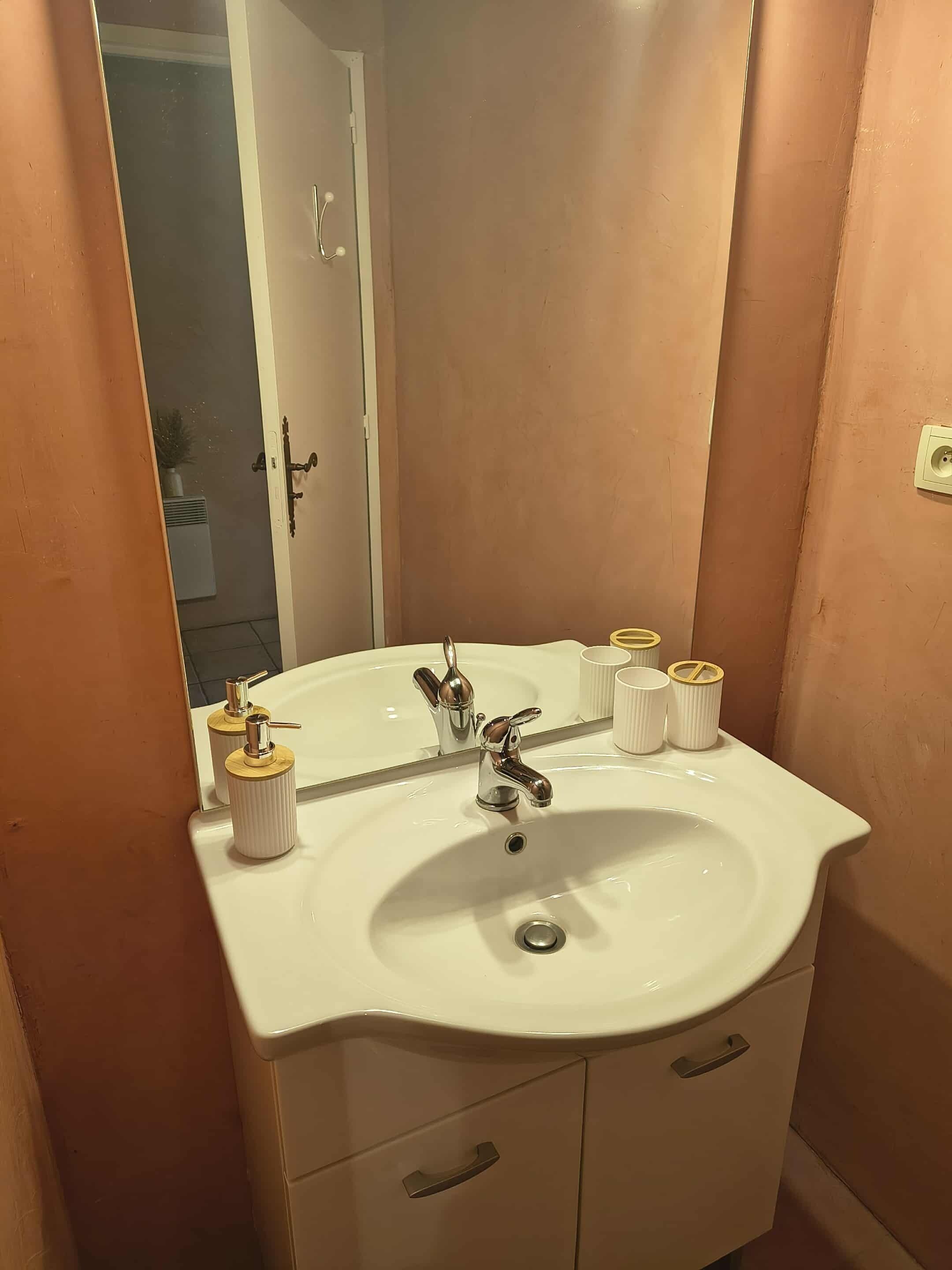 Une salle de bain avec un lavabo blanc, deux robinets, un miroir et trois articles de toilette rangés dans un porte-savon.