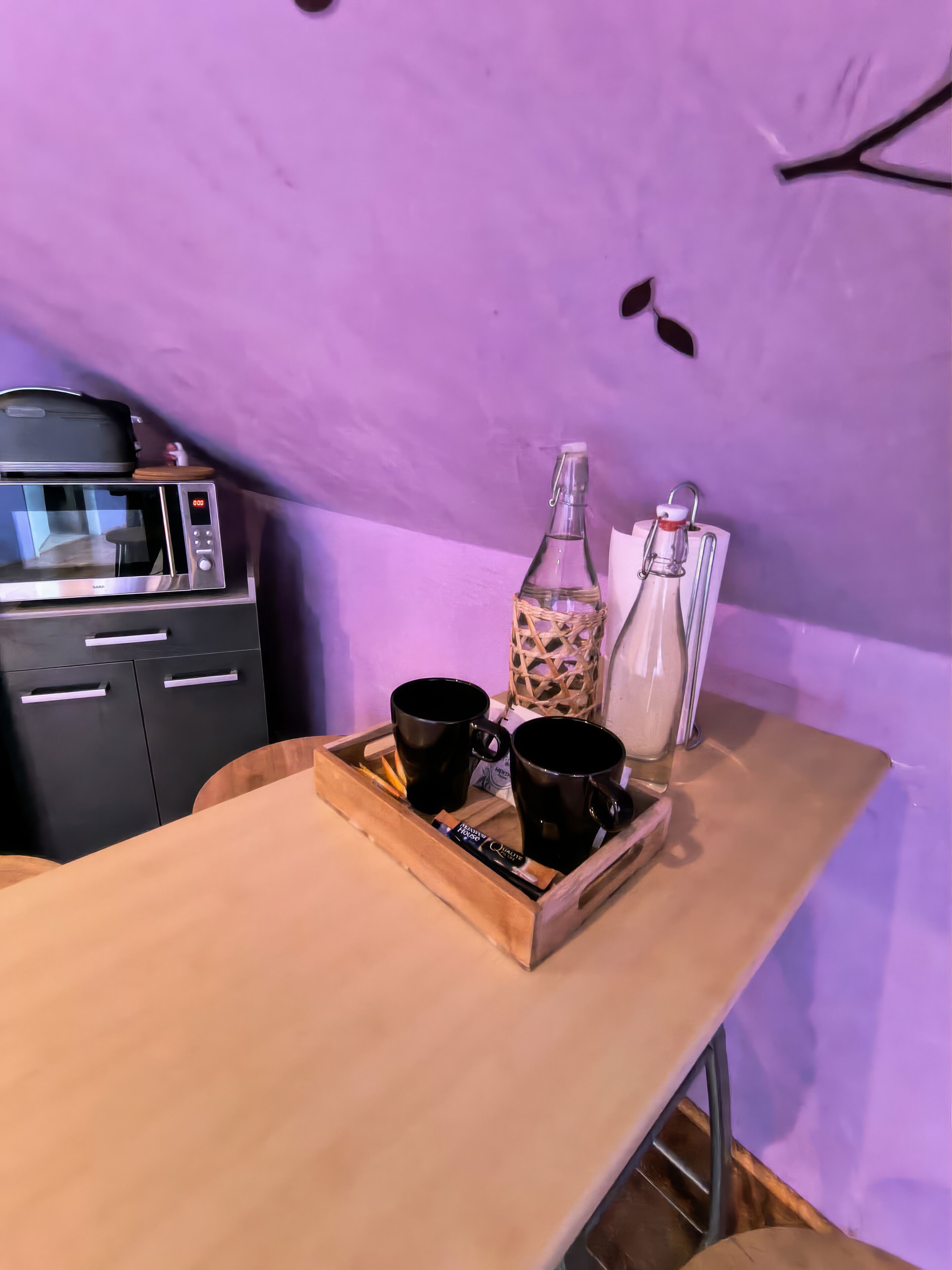 A wooden tray with two black mugs and a bottle on a table, a microwave, and a cabinet in a room with purple walls.