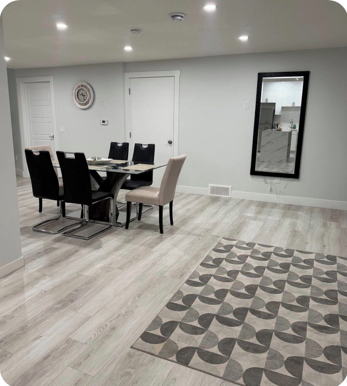 A dining area with a table and chairs, a clock on the wall, and a patterned rug.