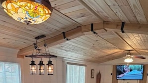 A spacious living area with a wooden ceiling, a dining table with bar stools, a sofa, and a television mounted on the wall.