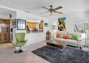 Ein modernes Wohnzimmer mit einem grünen Sessel, einem weißen Sofa mit bunten Kissen, einem schwarzen Teppich und einem Deckenventilator.